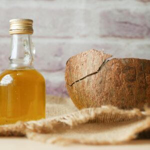 Bottle of coconut oil beside a cracked coconut shell on burlap fabric indoors.